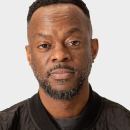 A man with short hair and a beard, wearing a black quilted jacket, looks directly at the camera against a plain light background.