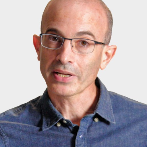 A middle-aged person with glasses and a receding hairline, wearing a blue button-up shirt, is speaking against a plain light background.