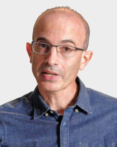 A middle-aged person with glasses and a receding hairline, wearing a blue button-up shirt, is speaking against a plain light background.