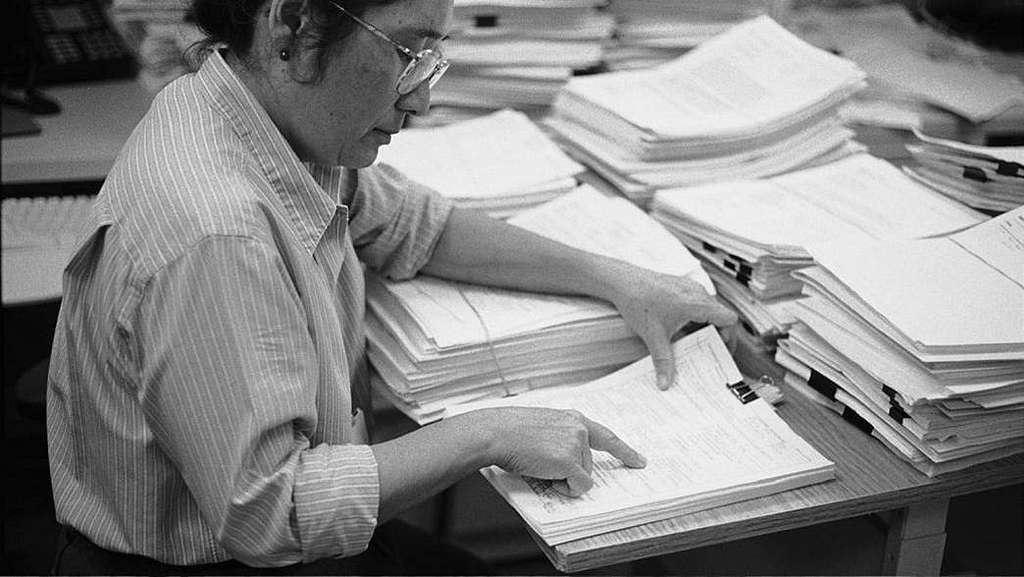 A woman sits at a desk covered with tall stacks of papers, reviewing and pointing to documents as she conducts a purpose-driven peer review in a busy office setting.