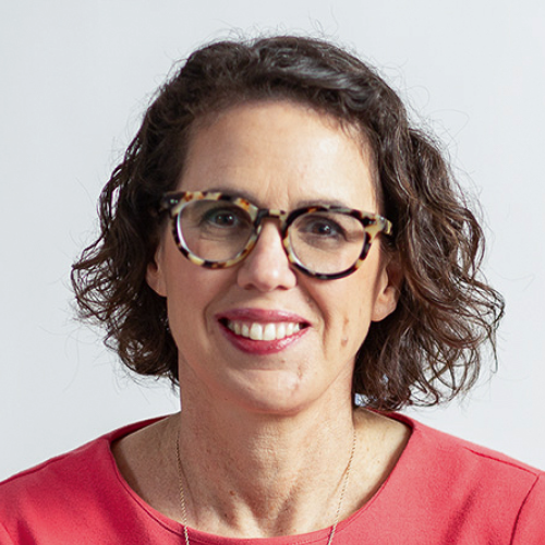 A woman with short curly brown hair, wearing tortoiseshell glasses and a coral top, smiles at the camera against a plain light background.