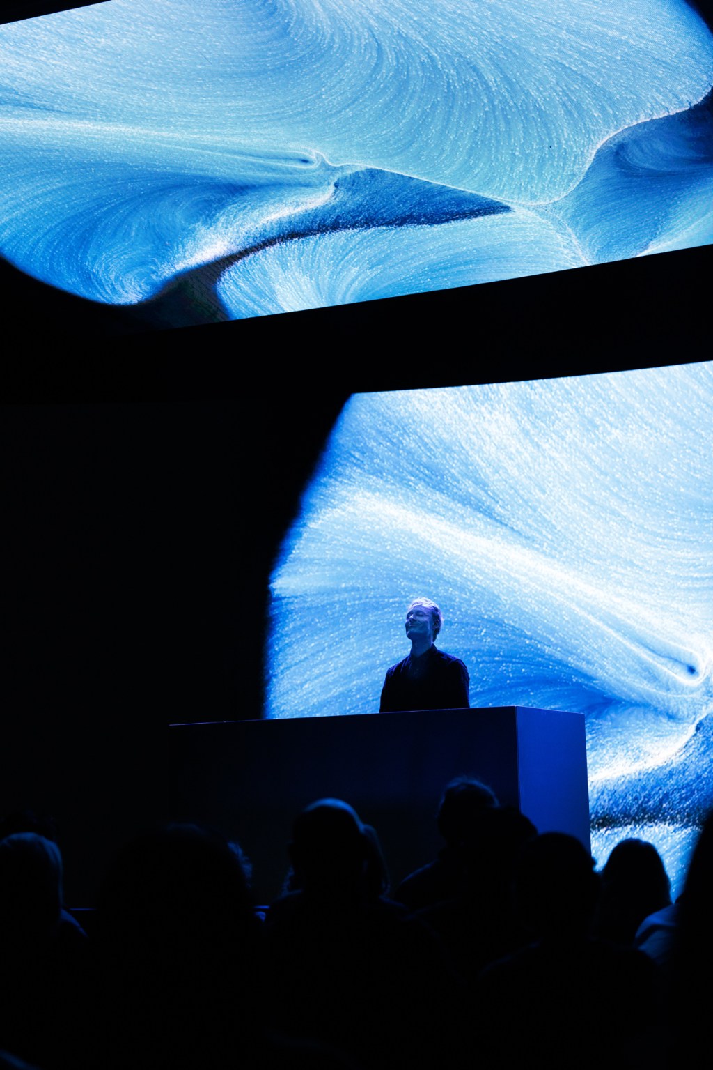 A person stands at a podium on a dark stage with large, abstract blue swirling visuals displayed on screens behind them.
