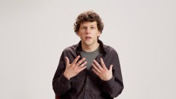 A person with curly brown hair wearing a black shirt and gray t-shirt sits against a plain white background, gesturing with both hands raised near their chest.