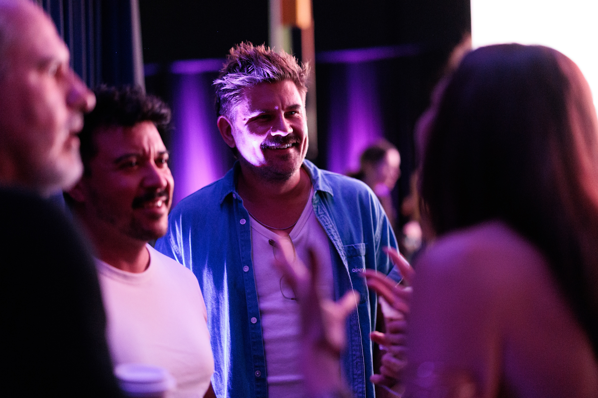 Three people stand talking in a dimly lit indoor setting with purple lighting, while a fourth person gestures in the foreground.