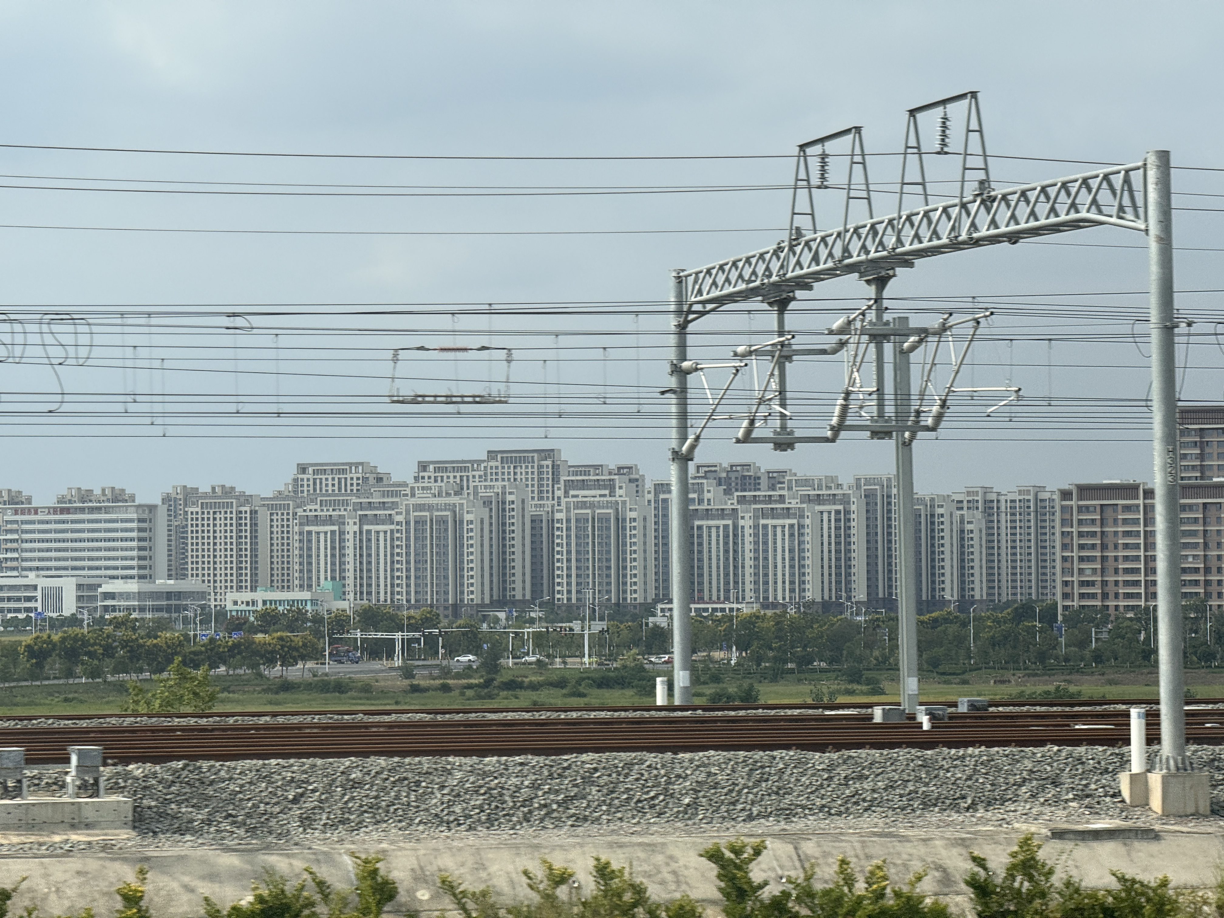 High-rise residential buildings in China stand in the background, with railway tracks and overhead electrical wires in the foreground under a cloudy sky.