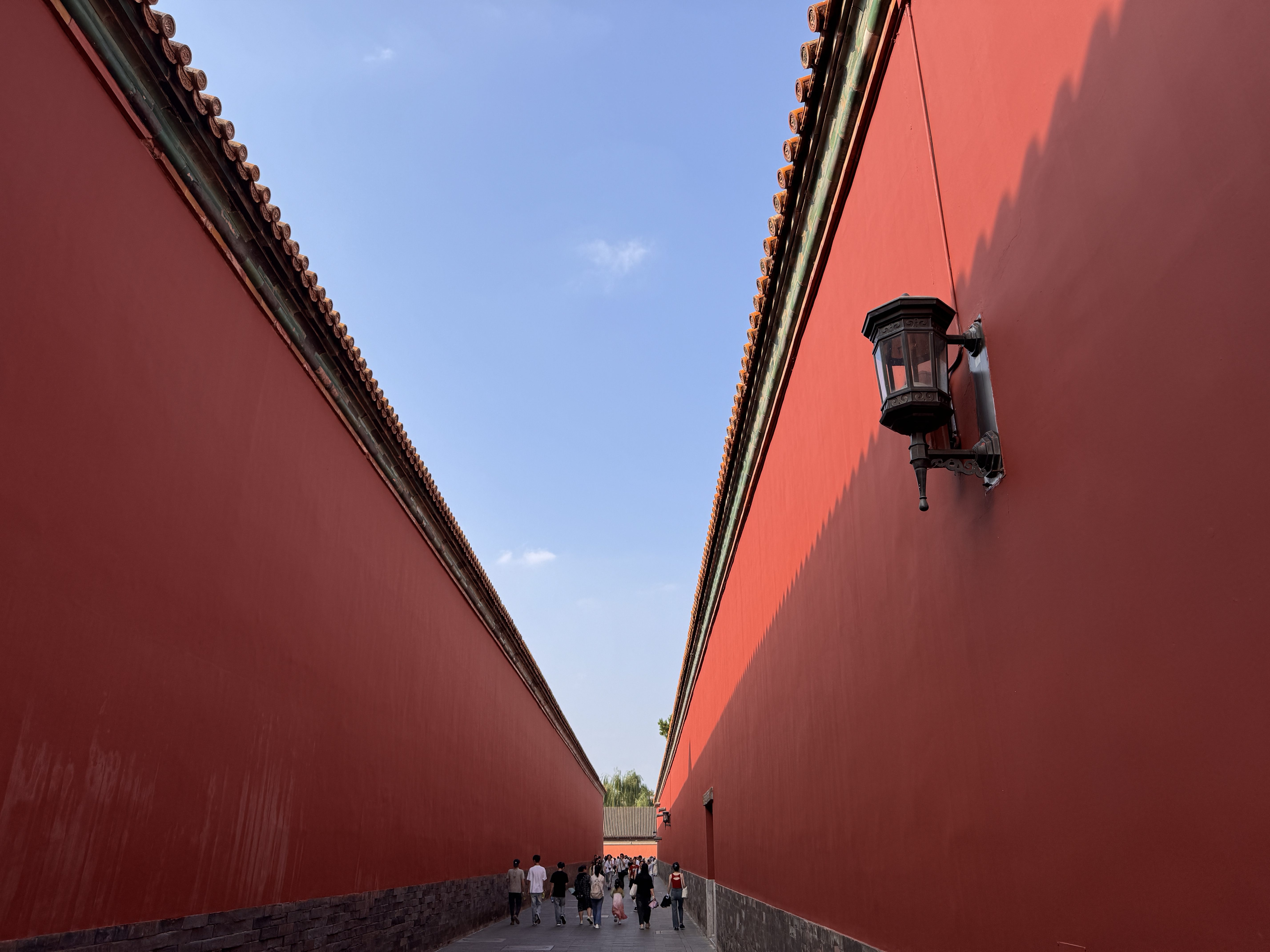 A group of people walk between two tall red walls in China, with a lantern hanging on the right wall under a blue sky.